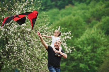 Spending weekend together. Running with red kite. Child sitting on the man's shoulders. Having fun