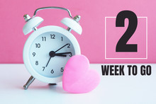 A Small White Alarm Clock And A Pink Heart Figurine. The Inscription "2 Week To Go"...