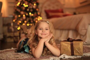 A cute girl with blond hair with a gift in her hands is waiting for the holiday against the background of the Christmas tree in the lights.