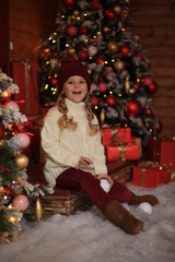 Wall Mural - cute beautiful girl in a white sweater and red hat having fun and playing snowballs in winter on the street against the background of a wooden house. nearby are Christmas trees with lights