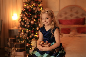 Wall Mural - A cute girl with blond hair with a gift in her hands is waiting for the holiday against the background of the Christmas tree in the lights.