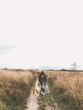© sonyachny - Stylish Hipster girl in yellow hat and coat walking with her golden dog in coat in autumn field among herbs. Woman in modern clothes walking with friend dog in countryside