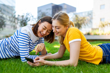 Wall Mural - smiling mother and daughter lying on grass in park looking at mobile phone