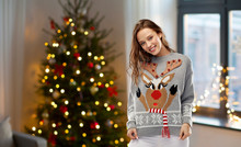 People And Holidays Concept - Happy Young Woman Wearing Ugly Sweater With Reindeer Pattern Over Home And Christmas Tree Lights On Background
