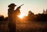 Fototapeta Sport - Silhouette of a hunter in a cowboy hat with a gun in his hands on a background of a beautiful sunset. The hunting period, the fall season is open, the search for prey.