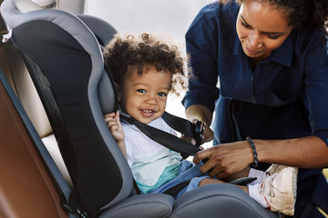 Side view of a happy little boy looking at camera while his mother buckling him in a car seat