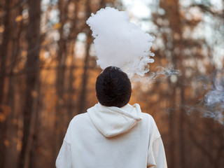 Wall Mural - A young man in a white hoodie exhales a cloud of steam smoke up against the background of an autumn Park