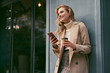 © Anton - Attractive cheerful blond girl in stylish trench coat with coffee to go happily using cellphone outdoor