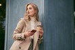© Anton - Attractive casual blond girl in stylish trench coat with coffee to go and cellphone thoughtfully standing outdoor