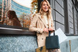 © Anton - Pretty cheerful stylish blond girl in beige coat with shopping bags happily looking in camera on city street