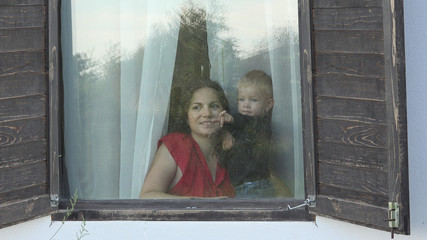 Mother and baby child looking out the window with shutters, outdoor view