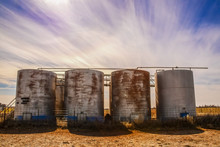 Old Rusty Holding Tanks With Shadows At Oil Well Location On Flat Farm Land Under Dramatic Sky At Golden Hour