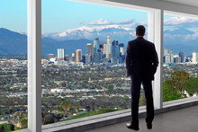 Businessman In An Office Looking At The View Of Downtown Los Angeles. The Man Looks Like A Boss Or A Regional Manager Working In California. The Background Is Snowy San Gabriel Mountains In Winter.
