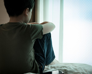 Cyberbullying concept. Young Asian preteen/teenage boy sit alone on the bed absent-minded hugging knees with his smartphone on his side. Stressed, frustrated, overwhelmed with online bullying.