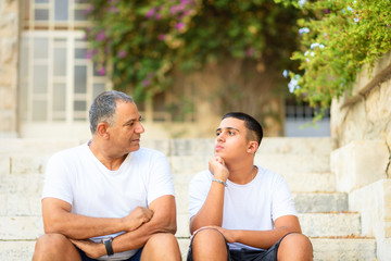 Wall Mural - Teenager son and senior father sitting on stairs outdoors at home, talking.