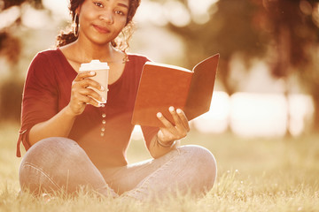 Friendly girl satisfied with weather, coffee and new book. Cheerful african american woman in the park at summertime