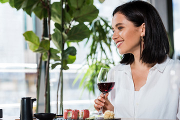 Wall Mural - happy woman holding glass with red wine near tasty meal in sushi bar