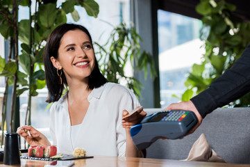 Wall Mural - cropped view of waiter holding credit card reader near smiling woman paying with smartphone