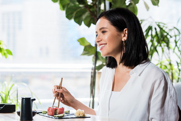 Wall Mural - young happy woman holding chopsticks near delicious sushi in restaurant