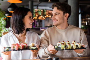 happy man and cheerful woman smiling while holding plates with sushi