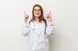 © Asier - Young doctor woman against a white wall indicates with both fore fingers up showing a blank space.
