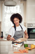 © nenadaksic - Smiling mixed race woman in apron putting chopped mushrooms in bowl with vegetables. Kitchen interior.
