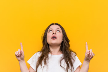 Wall Mural - Young curvy plus size woman pointing upside with opened mouth.