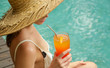 Girl in a big straw hat sitting at the pool edge and holding a cocktail glass, summertime vacation leisure activity