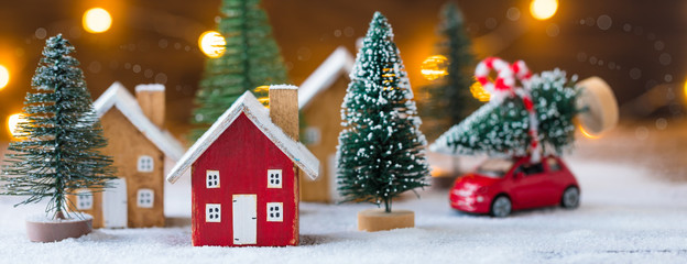 miniature wooden houses and small red car with fir tree on the snow over blurred christmas decoratio