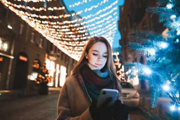 Girl stands on the street near the Christmas tree and on the background of Christmas lights and uses a smartphone. Attractive girl uses smartphone on the street with Christmas decorations. New Year