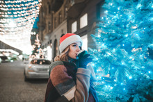 Portrait Of A Beautiful Girl In A Hat Of Santa Claus Stands On A Christmas-decorated Street Near A Christmas Tree, Looking Away And Drinking Coffee. New Year Portrait Of A Cute Girl. Christmas Concept