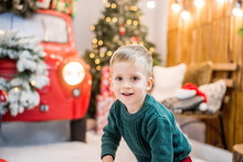 Portrait Of A Little Happy Boy Who Plays With Red-white Boxes Of Gifts, Builds Towers Against The Background Of A Car With A Wreath, Bokeh, Christmas Trees With Garlands Of Lights. New Year Copy Space