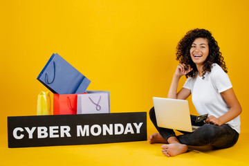 happy black girl with colorful shopping bags and cyber monday sign sitting with laptop and credit card isolated over yellow