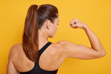 Horizontal Shot Of Attractive Fitness Woman Posing Backwards Isolated Over Yellow Studio Background. Trained Female Body, Girl Showing Her Biceps, Lady With Ponitail Wearing Black Sport Top.