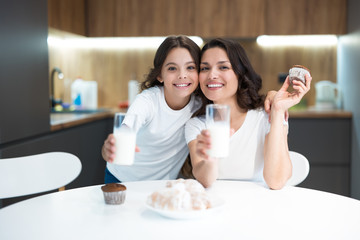 beautiful young mother with her cute teenager daughter eating cupcakes and croissants and drinking milk and kefir for breakfast in the kitchen looking happy