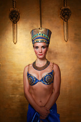 Wall Mural - Beautiful thin Egyptian woman with pronounced cheekbones in a huge gold headdress without hair in a blue swimsuit on the background in the Studio