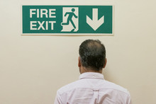  Man Seen Walking Into A Solid Wall, Below A Fire Exit Sign Located In An Office Complex. The Actual Fire Exit Is Down The Stairs, This Being A Funny Concept Image.