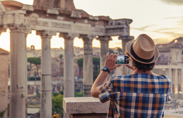 Young man tourist backpacker taking pictures with smartphone at Roman Forum at sunrise. Historical imperial Foro Romano in Rome, Italy from panoramic point of view.