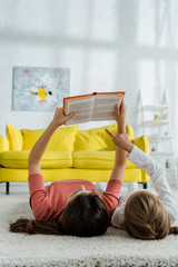 Wall Mural - child pointing with finger at book while lying on carpet with babysitter