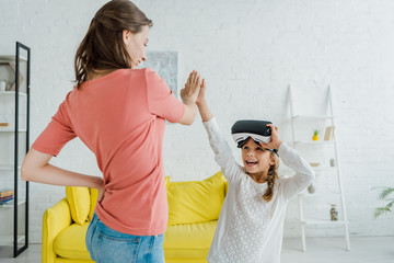 Wall Mural - cheerful kid in virtual reality headset giving high five to babysitter at home