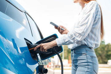 Young adult woman charging electric car, using smartphone