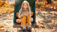 Little Girl Sitting On Ripe Pumpkin Against Wheelbarrow With Autumn Harvest