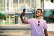 Happy young African bearded man with Afro hair taking selfie in city