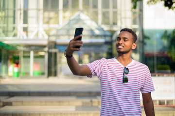 Wall Mural - Happy young African bearded man with Afro hair taking selfie in city