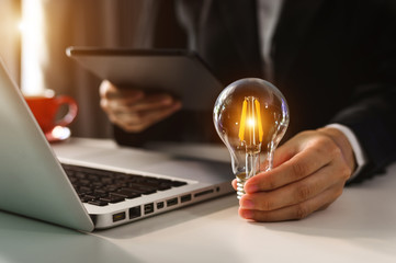 business hand holding lightbulb with using laptop computer and money stack in office. idea saving energy and accounting finance concept  in morning light