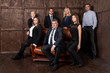 © spritnyuk - A team of businessmen stands and sits on a brown sofa against a dark wooden wall
