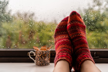 Woman Feet In Warm Wool Socks Next To Colourful Mug With Fall Leaves Against A Rainy Widow Background. Autumn Rain And Cold Weather Concept.