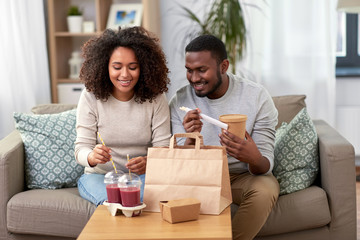 Wall Mural - eating and people concept - happy african american couple with takeaway food and drinks at home