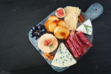 Appetizer platter with assorted cheeses and deli meat. Overhead view on a dark stone background.