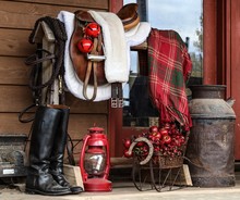 Portrait Of A Black Great Dane Resting On A Farmhouse Porch Beside A Saddle Rack With English Tack Including Saddle, Bridle, Riding Boots Decorated For Christmas- Bells, Lantern And Plaid Blanke